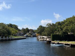 Canal lined with sea walls on either side with boats docked at piers, lots of trees, lawns and a view of a mansion in the distance.