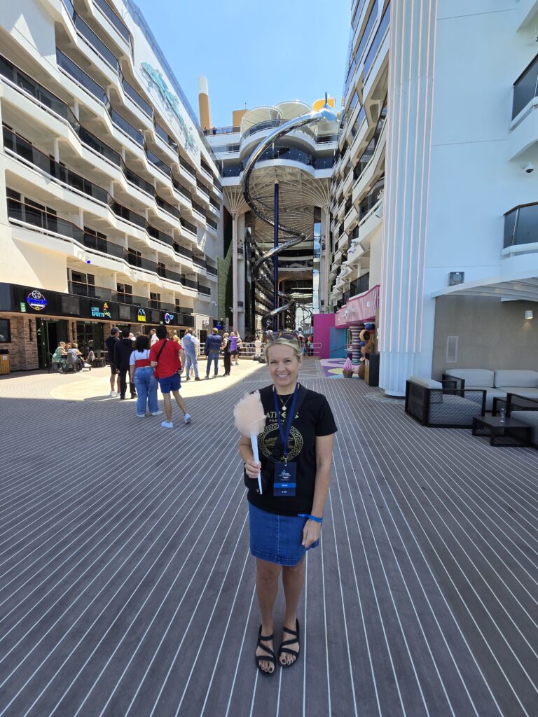 A pretty blond writer stands in the shade of a megaship's superstructure, smiling and holding a cardboard cone that's engulfed by a large, orange cloud of cotton candy aboard MSC America