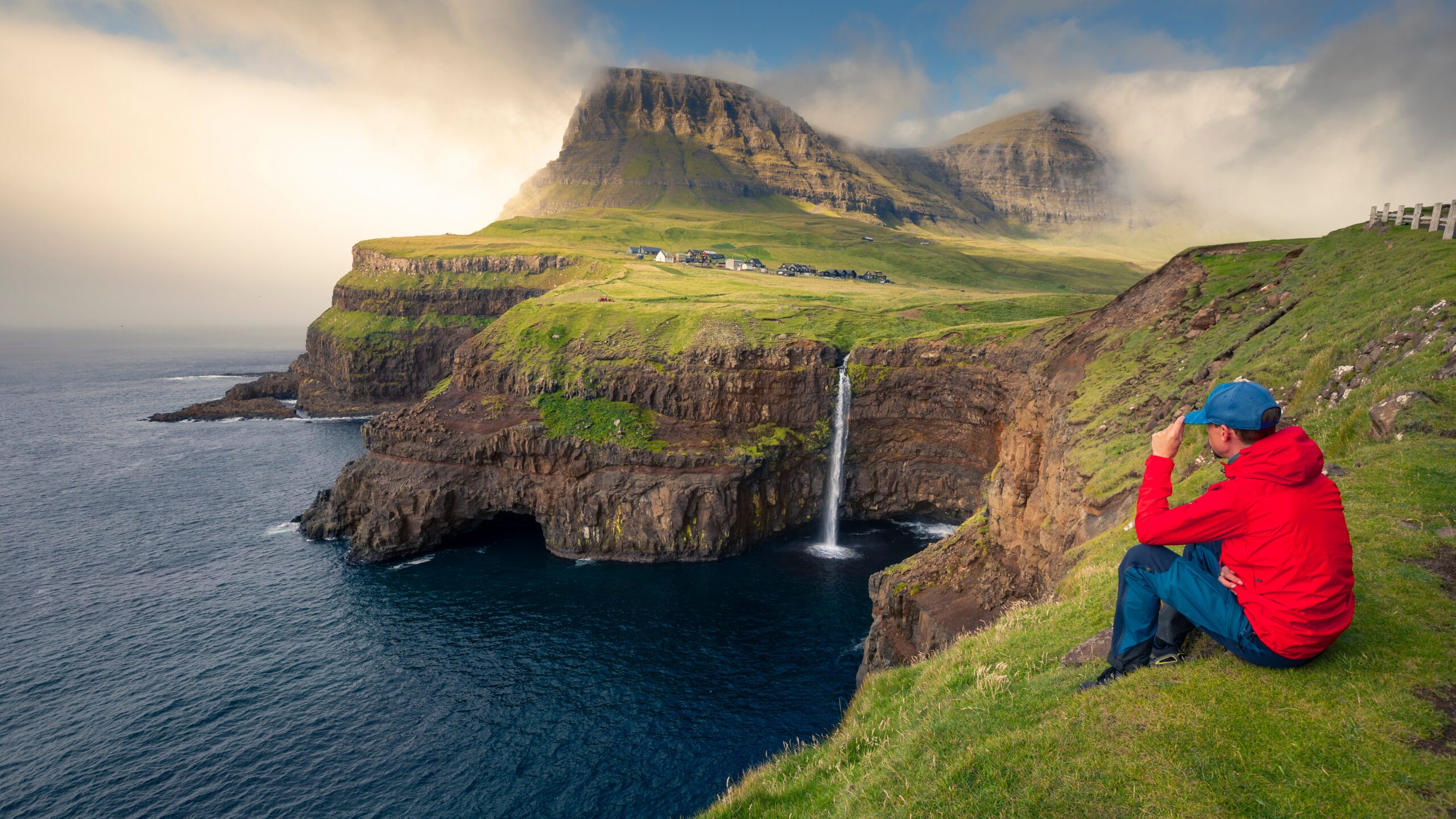Man sits at Múlafossur waterfall with Gásadalur village on Vagar island, Faroe Islands